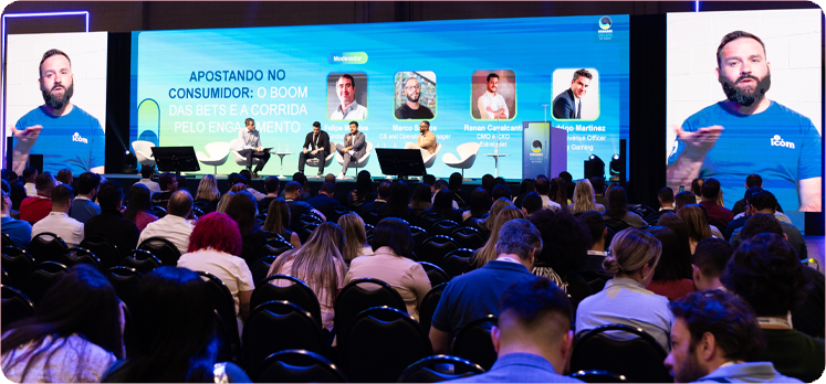 Evento com painel sobre a indústria de apostas, apresentando quatro pessoas em uma mesa de discussão, com uma multidão de ouvintes ao fundo e um grande telão exibindo o título do painel sobre engajamento do consumidor.