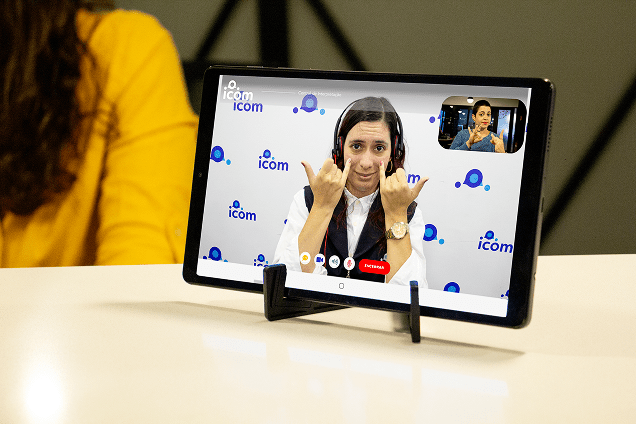 Uma tablet mostrando uma chamada de vídeo com uma pessoa usando fones de ouvido e fazendo sinais com as mãos em frente a um fundo com o logotipo da iCom. A pessoa à esquerda é parcialmente visível, com uma blusa amarela.