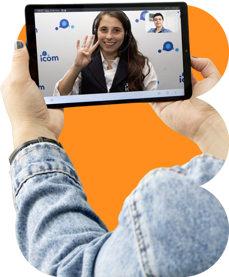 Uma pessoa segurando um tablet mostrando uma videochamada com uma mulher sorridente usando fones de ouvido, que faz um gesto de 'número quatro'. O fundo da videochamada apresenta o logo 'icom' em uma parede branca.