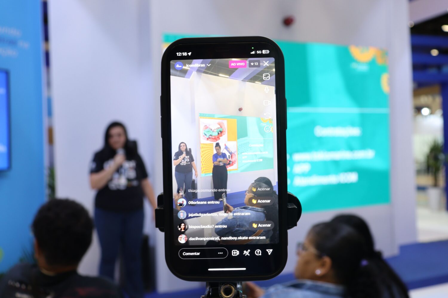 Uma pessoa falando ao microfone em um evento, visível através da tela de um smartphone em uma transmissão ao vivo. Ao fundo, outra pessoa está ao lado e um telão exibe informações em cores vibrantes.