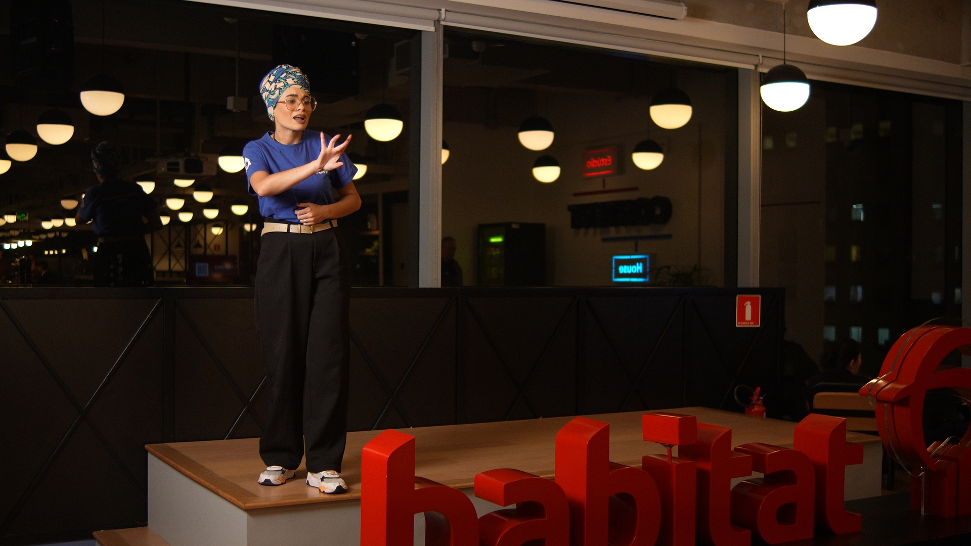 Uma mulher com turbante, óculos e camiseta azul está em pé sobre um palco com letras grandes vermelhas que dizem 'habitat'. O ambiente é moderno e bem iluminado, com mesas e pessoas ao fundo.