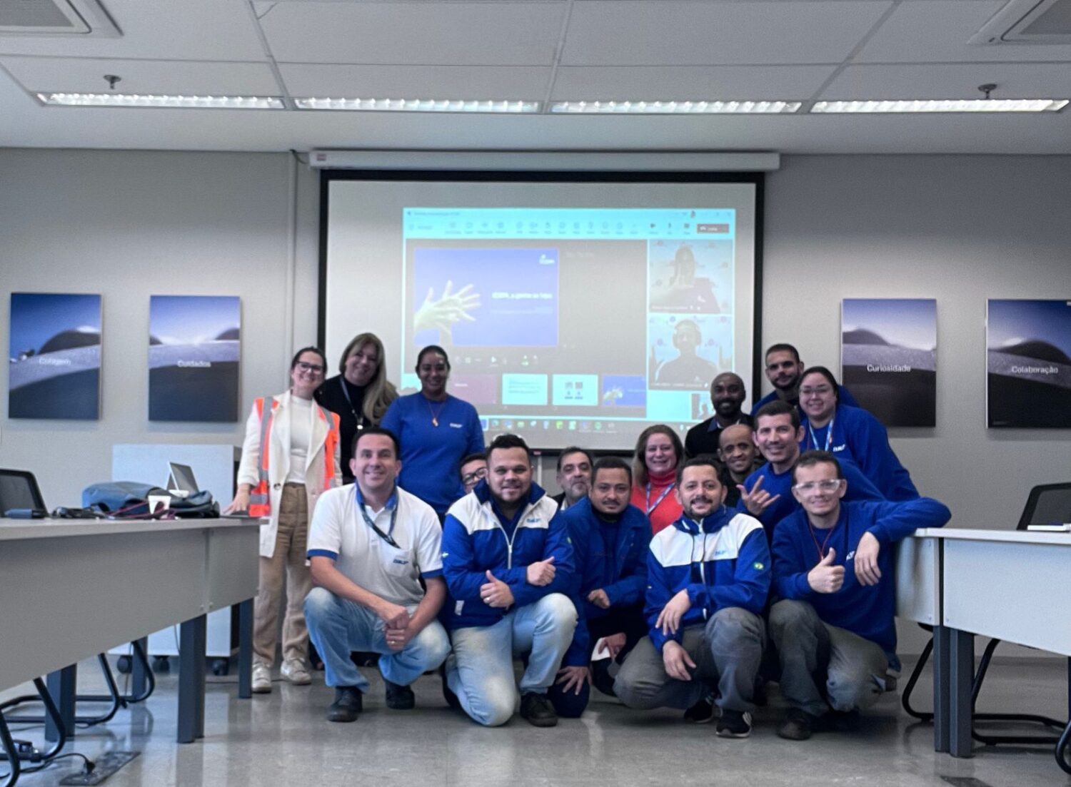 Um grupo de pessoas sorrindo em uma sala de reuniões, com várias pessoas vestindo roupas azuis. Ao fundo, uma apresentação em um projetor e imagens nas paredes. Uma mulher à frente usa um colete laranja.