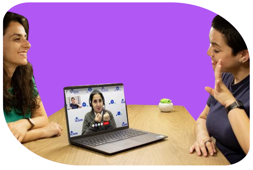 Duas mulheres sentadas em uma mesa, uma delas olhando para uma laptop que exibe uma videoconferência, enquanto a outra mulher está interagindo de forma animada, com um fundo roxo e um pequeno objeto decorativo na mesa.